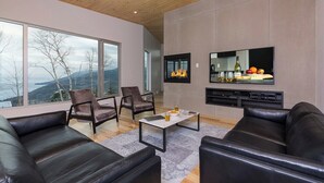A modern living room with leather sofas, a fireplace, a flat-screen TV, and a large window overlooking a mountainous landscape. - Le Rupin 77: River, Mountain Views & Hot Tub (Charlevoix)