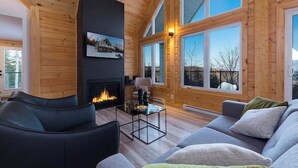 Interior acogedor de una cabaña con chimenea, paredes de madera, ventanas amplias y un sofá moderno.