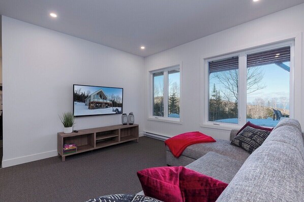 A modern living room with a grey sofa, a red throw blanket, and a large window offering a view of trees and a building.