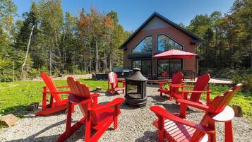 A modern lodge with a large glass facade, surrounded by a gravel patio with red Adirondack chairs and a fire pit.