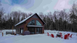 Una cabaña con un ventanal grande, un porche cubierto y sillas Adirondack rojas en la nieve.