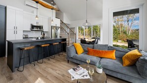 A modern kitchen with white cabinets, a black countertop, and a stainless steel refrigerator. There is a dining area with a gray sofa and yellow cushions. - Nordik 126: Private Hot Tub, Covered Terrace & BBQ (Les Éboulements)
