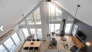 A spacious loft-style room with large windows, a dining area, a seating area, and a TV. - Nordik 126: Private Hot Tub, Covered Terrace & BBQ (Les Éboulements)