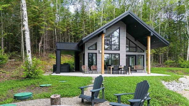 A modern cabin with a large deck, outdoor seating, and a fire pit. - Nordik 126: Private Hot Tub, Covered Terrace & BBQ (Les Éboulements)