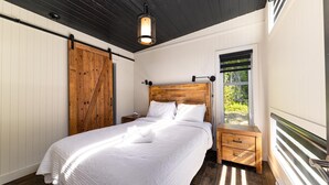 Ein Schlafzimmer mit einem hölzernen Kopfteil, einem Bett mit weißer Bettwäsche, einem Nachttisch, einer Holzkommode, einer Schiebetür im Bauernhausstil und einem Fenster mit Blick auf Grünflächen.