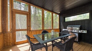 Eine Holzhütte mit Essbereich, inklusive Glastisch, Korbstühle, Grill und großen Fenstern mit Blick auf den Wald.