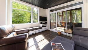 Ein modernes Wohnzimmer mit großem Fenster, Flachbildfernseher und einer Sitzecke mit Sofa und Hocker.