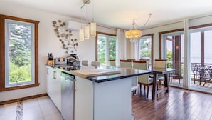 A modern kitchen with a central island, large windows, and a dining area with a view. - 14B Condo: Mountain & River Views, Terrace & BBQ (Petite-Rivière-Saint-François)