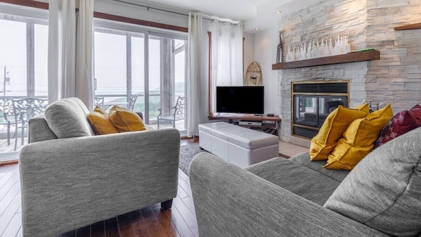 A modern living room with a stone fireplace, grey sofas with yellow cushions, a flat-screen TV, and large windows offering a view of the outdoors. - 14B Condo: Mountain & River Views, Terrace & BBQ (Petite-Rivière-Saint-François)
