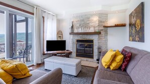 A living room with a stone fireplace, a flat-screen TV, and a sofa with yellow and red cushions. - 14B Condo: Mountain & River Views, Terrace & BBQ (Petite-Rivière-Saint-François)