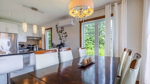 A modern kitchen with a central island, pendant lights, and a dining area with a wooden table and white chairs. - 14B Condo: Mountain & River Views, Terrace & BBQ (Petite-Rivière-Saint-François)