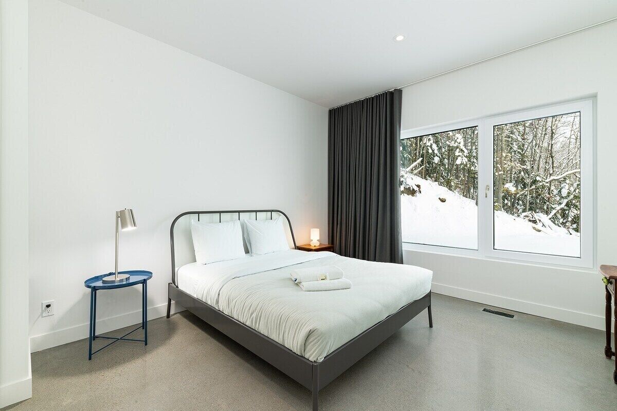 A modern bedroom with a bed, a side table, a lamp, and a large window with a view of a snowy landscape.