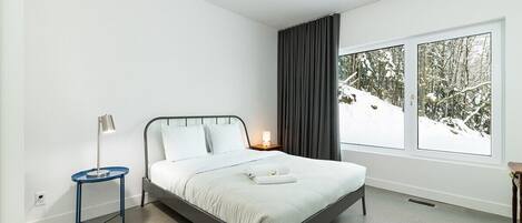 A modern bedroom with a bed, a side table, a lamp, and a large window with a view of a snowy landscape.