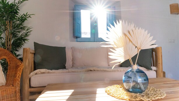 Una mesa de madera con un florero de vidrio y plantas secas, una silla de mimbre, un sofá con cojines y una ventana con luz solar entrando.