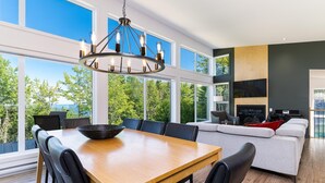 Un comedor moderno con una mesa y sillas de madera, una araña y ventanas amplias que ofrecen vistas al exterior.