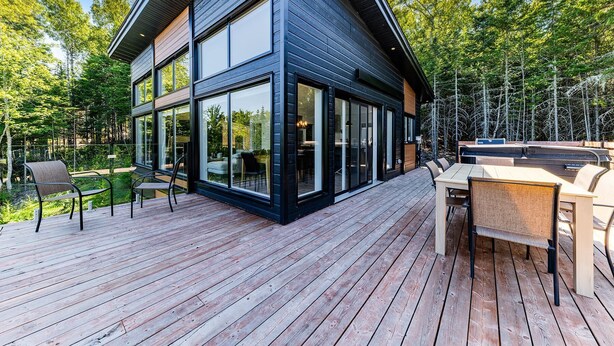 Una casa moderna con amplios ventanales, una terraza de madera y mobiliario exterior.