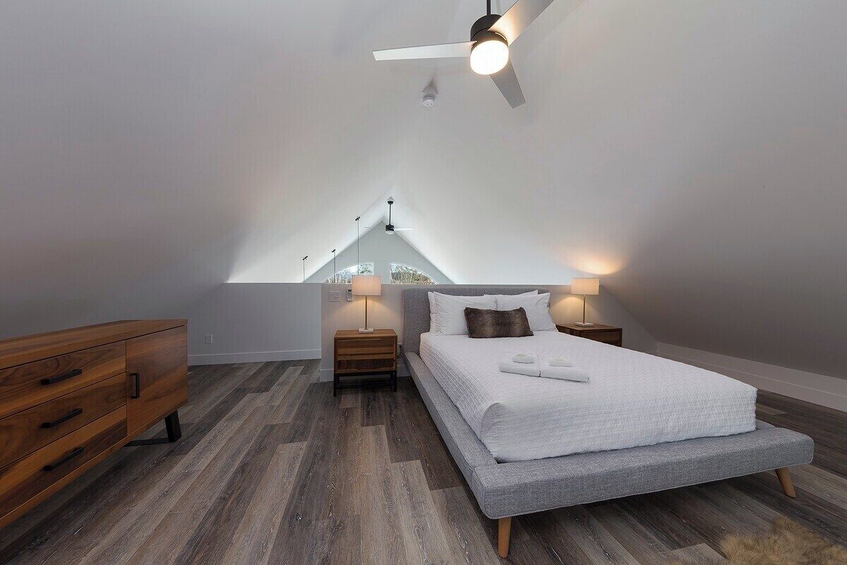 Ein modernes Schlafzimmer mit schrägverlaufender Decke, einem Bett mit weißer Bettwäsche, zwei Nachttischlampen, einer hölzernen Kommode und einem Deckenventilator.