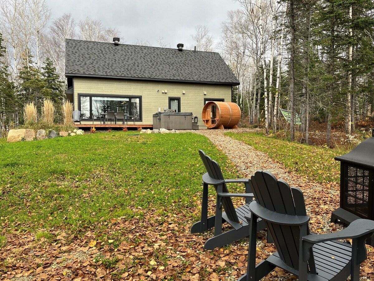 Una casa moderna con un gran ventanal, un barril de madera y dos sillas Adirondack en el jardín.