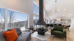 A modern living room with a fireplace, large windows, and a dining area. - Le Chaleureux 61: River & Mountain Views (Petite-Rivière-Saint-François)