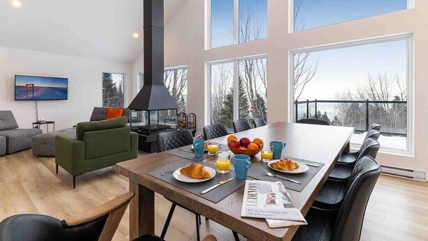 A modern living room with a dining area, large windows, and a fireplace. - Le Chaleureux 61: River & Mountain Views (Petite-Rivière-Saint-François)