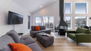 A modern living room with a large sofa, ottoman, and a green armchair. There is a fireplace and a flat-screen TV mounted on the wall. - Le Chaleureux 61: River & Mountain Views (Petite-Rivière-Saint-François)