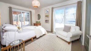 A bedroom with a bed, a chair, a bedside table, a lamp, a rug, and a large window with curtains. - Chez Annie 215: Panoramic Views & Hot Tub (Charlevoix)