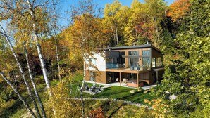 A modern two-story house with a glass facade, surrounded by a well-maintained garden and trees. - Chez Annie 215: Panoramic Views & Hot Tub (Charlevoix)