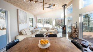 A modern living room with a wooden ceiling, large windows, a fireplace, and a dining table with a fruit bowl. - Chez Annie 215: Panoramic Views & Hot Tub (Charlevoix)