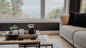 A cozy living room with a large window, a wooden coffee table with a tray, a sofa, and a black lamp. - Refuge 2: Hot Tub, River Views & Pet Friendly (Les Éboulements)