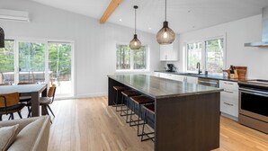 A modern kitchen with a central island, bar stools, pendant lighting, and large windows. - Refuge 2: Hot Tub, River Views & Pet Friendly (Les Éboulements)
