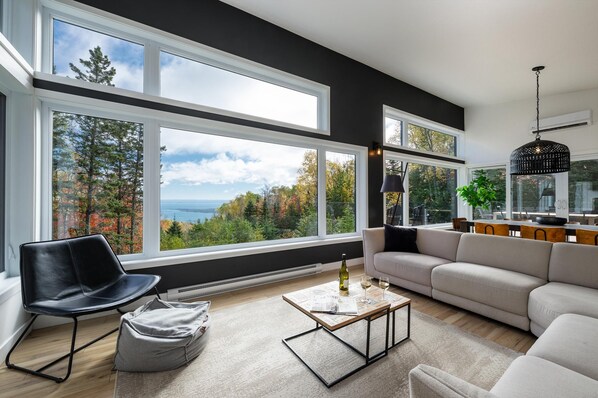 A modern living room with large windows, a sofa, a coffee table, and a dining area. - Refuge 2: Hot Tub, River Views & Pet Friendly (Les Éboulements)