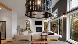 A modern living room with a sofa, a dining table with apples, a fireplace, and large windows. - Refuge 2: Hot Tub, River Views & Pet Friendly (Les Éboulements)