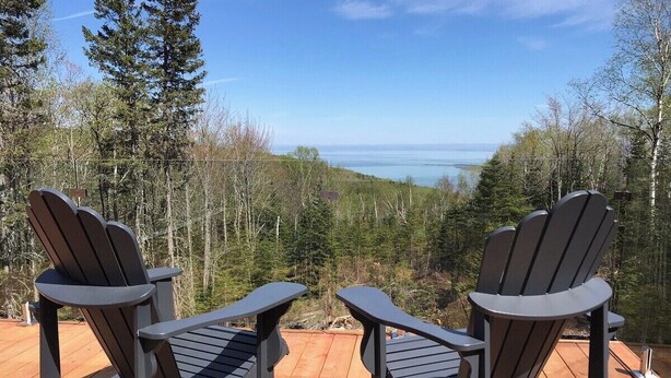 Two Adirondack chairs on a wooden deck with a view of a forest and a body of water. - Refuge 2: Hot Tub, River Views & Pet Friendly (Les Éboulements)