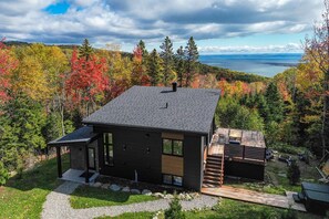 A modern two-story cabin with a large deck and a view of a lake surrounded by autumn foliage.
