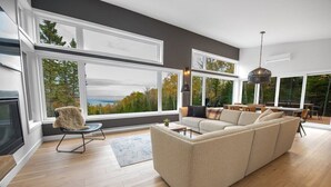 A modern living room with a large beige sectional sofa, a round coffee table, and a chair. There are large windows offering a view of trees and a cloudy sky. - Refuge 2: Hot Tub, River Views & Pet Friendly (Les Éboulements)