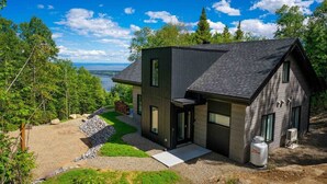 Una casa moderna de dos pisos con fachada oscura, ventanas grandes y vistas a un lago.