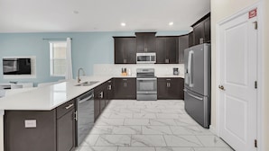 Uma cozinha moderna com armários escuros, bancada branca, eletrodomésticos de aço inoxidável e piso revestido de azulejos.