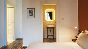 Hotellihuone, jossa on sänky, yöpöytä lampun kanssa, pieni kylpyhuone peilillä ja pesualtaalla sekä ovi, joka johtaa toiseen huoneeseen.