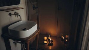 Kylpyhuoneessa on valkoinen pesuallas, peili ja sen yläpuolella hylly. Lattialla on kynttilöitä, ja taustalla näkyy suihku.