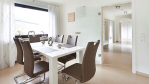 A modern dining area with a white table and brown chairs, a window with curtains, and a hallway leading to another room. - Rheinflair Comfortable Holiday Residence (Kamp-Bornhofen)