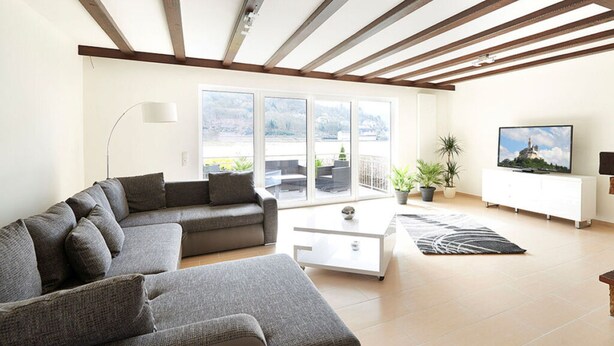 A modern living room with a sectional sofa, a white coffee table, a flat-screen TV, and a balcony with potted plants. - Rheinflair Comfortable Holiday Residence (Kamp-Bornhofen)