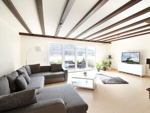 Un salon moderne avec un canapé d’angle, une table basse blanche, un téléviseur à écran plat et un balcon agrémenté de plantes en pot.