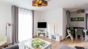 Ein modernes Wohnzimmer mit einem Flachbildfernseher, einem Glastisch, grauen Sofas, einem Essbereich mit einem weißen Tisch und Stühlen sowie einem Gemälde an der Wand.
