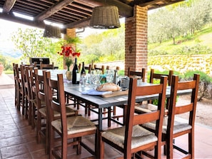 An outdoor dining area with a brick column, wooden chairs, a table set for dining, and a view of a garden. - Villa in Umbria With Private Pool & Valley Views (Perugia)