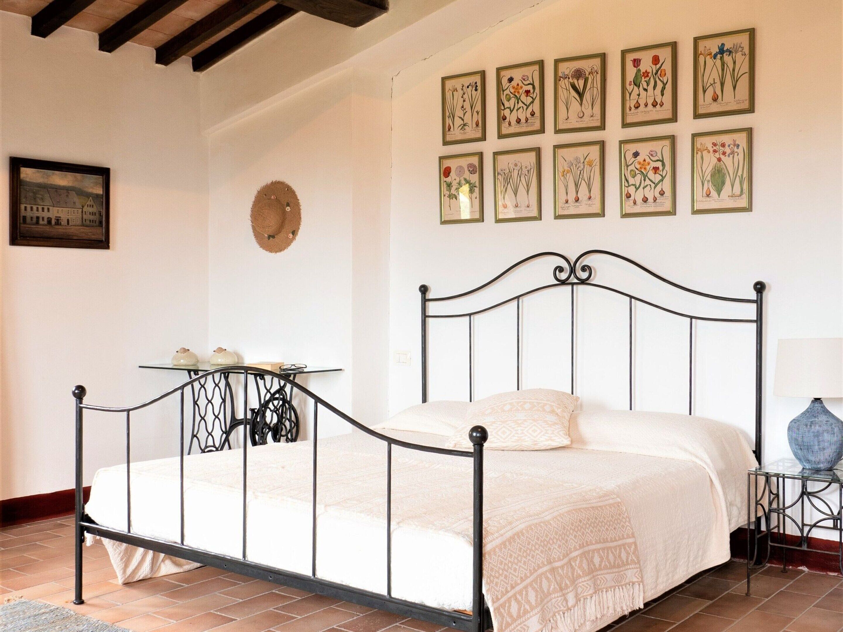 Un dormitorio con estructura de cama metálica, una mesita de noche, una lámpara e ilustraciones botánicas enmarcadas en la pared.