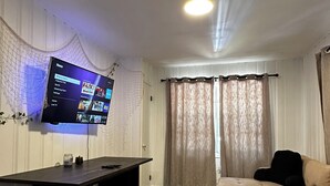 A living room with a flat-screen TV mounted on the wall, a dark wooden table, a beige sofa, and a woven rug.