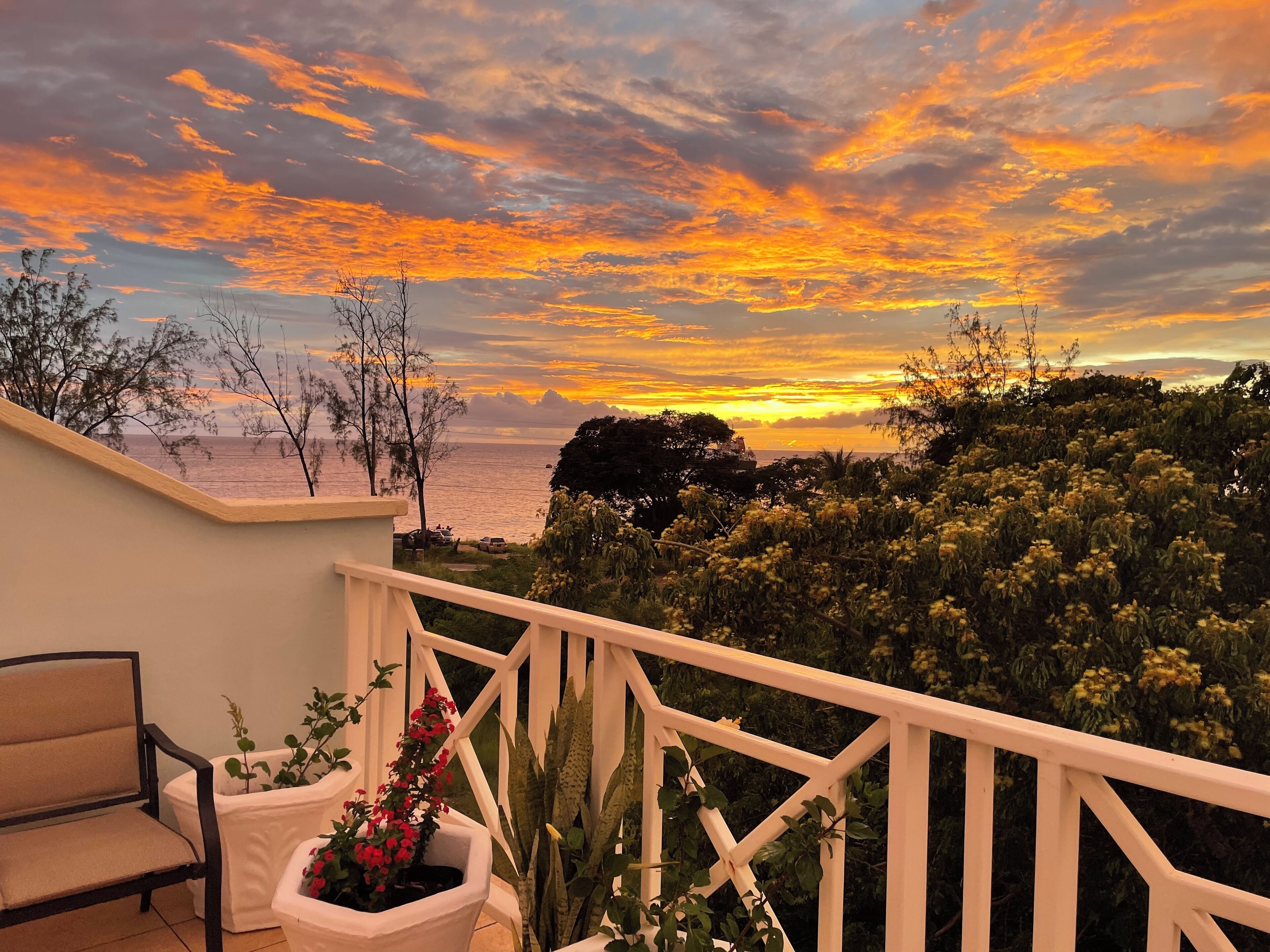 Un balcon avec vue sur le coucher de soleil sur la mer, une chaise et des plantes en pot.