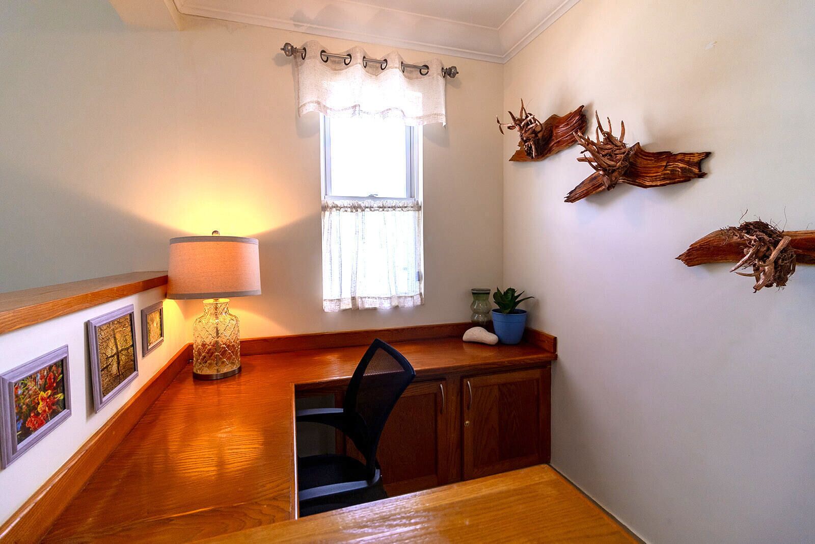 A small, cozy room with a wooden desk, a chair, a lamp, a window with curtains, and wall-mounted decor.