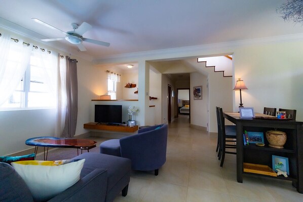 A living room with a TV, sofa, chairs, a dining table with chairs, a staircase, and a ceiling fan.