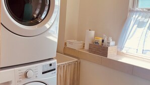 Un espace buanderie comprenant deux machines à laver superposées, une fenêtre avec vue et une étagère contenant des produits de lessive.